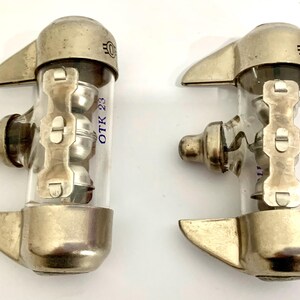 Puede incluir: Dos v&aacute;lvulas de vac&iacute;o vintage con componentes de metal y vidrio. Cada v&aacute;lvula tiene una base de metal plateado, un cilindro de vidrio transparente y un soporte de metal. El texto "OTK 23" est&aacute; impreso en el vidrio. Las v&aacute;lvulas tienen un dise&ntilde;o industrial &uacute;nico.