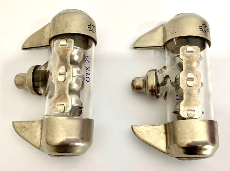 Puede incluir: Dos v&aacute;lvulas de vac&iacute;o vintage con componentes de metal y vidrio. Cada v&aacute;lvula tiene una base de metal plateado, un cilindro de vidrio transparente y un soporte de metal. El texto "OTK 23" est&aacute; impreso en el vidrio. Las v&aacute;lvulas tienen un dise&ntilde;o industrial &uacute;nico.