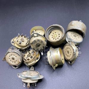 May include: A collection of ten vintage electronic vacuum tube sockets featuring circular ceramic bases with metallic outer shells and multiple protruding metal pins. The components exhibit varying degrees of oxidation and wear on the metal surfaces.