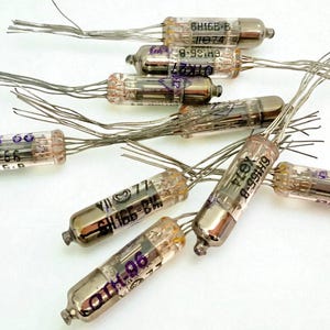 Puede incluir: Una colección de válvulas de vacío vintage con cuerpos de vidrio transparente y bases de metal plateado. Cada válvula tiene múltiples cables y texto impreso, incluyendo números de modelo como "6H165-B" y "OTK27".