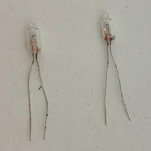 Könnte beinhalten: Zwei kleine, klare Glasglühbirnen mit silbernen Drähten. Die Glühbirnen haben eine kleine, zylindrische Form mit kupferfarbenen Filamenten im Inneren. Die Drähte sind leicht gebogen, vor einem hellen Hintergrund.