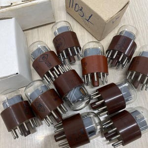 May include: A collection of vintage vacuum tubes with brown bases and clear glass bulbs. The tubes have multiple pins and are arranged with two cardboard boxes. Some tubes have printed markings. The boxes have handwritten text.