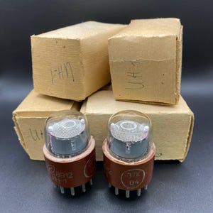 May include: Two vintage vacuum tubes with brown bases and clear glass tops, alongside four small, unmarked cardboard boxes. The tubes have visible internal components and are labeled with Cyrillic characters and numbers. The boxes have handwritten markings.