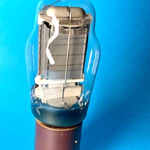Puede incluir: Una válvula de vacío vintage con una bombilla de vidrio transparente, una parte superior plateada y una base marrón con clavijas. Los componentes internos son visibles, incluyendo una estructura metálica gris y una cinta blanca. El tubo está sobre un fondo azul.