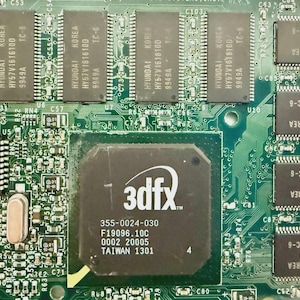 May include: Close-up of a green circuit board with various electronic components. The board features a central 3dfx chip and several Hyundai memory chips. Text on the 3dfx chip reads "3dfx" and other technical details. The board is filled with other small components.