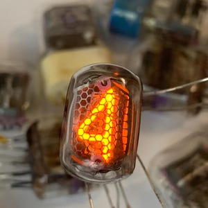 Może przedstawiać: Zbliżenie na zabytkową lampę Nixie wyświetlającą cyfrę 4 w jasnopomarańczowej poświacie. Lampa jest zamknięta w przezroczystym szkle, z wewnętrzną strukturą przypominającą plaster miodu. W tle widoczne są inne elementy elektroniczne.