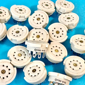 Może przedstawiać: Kolekcja zabytkowych białych ceramicznych gniazd lamp próżniowych z wieloma otworami na piny i centralnym złotym stykiem. Gniazda mają nadruk "ППк9 9-70". Tło jest jednolicie niebieskie.