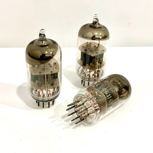 May include: Three vintage vacuum tubes with clear glass bodies and silver-coloured tops. The internal components are visible, including filaments and plates. The tubes have multiple pins at the base for electrical connections. One tube is lying on its side.