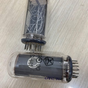 以下が含まれることがあります： 透明なガラスケースに入った2つのヴィンテージ真空管。1つのチューブは複雑な内部構造を示し、もう1つは印刷されたテキストと星のエンブレムを備えています。チューブには電気接続用の複数のピンが複数あります。