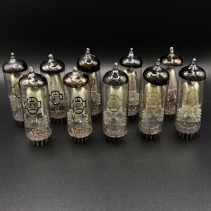 Può includere: Un gruppo di valvole termoioniche vintage con bulbi in vetro trasparente e cappucci metallici. Ogni valvola presenta componenti interni visibili e testo stampato. Le valvole sono disposte su una superficie scura, evidenziando il loro design.