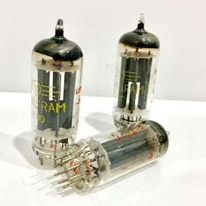 May include: Three vintage vacuum tubes with glass enclosures. The tubes have a clear glass body with internal components and metal pins. The top of each tube is dark, and some have visible text. The tubes are arranged on a white surface.