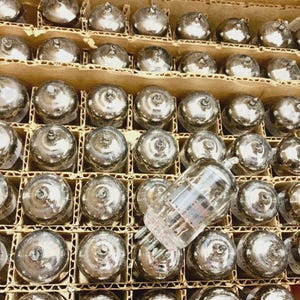May include: A collection of vintage vacuum tubes, each with a silver-coloured metal base and a glass enclosure. The tubes are neatly arranged in a cardboard box with individual compartments, showcasing a retro electronic component.