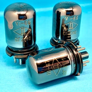 Pode incluir: Três válvulas de vácuo vintage com um acabamento metálico brilhante. Cada válvula tem as marcações "V-66", um logotipo em forma de diamante e "CF 201C". As válvulas estão dispostas sobre uma superfície azul clara.