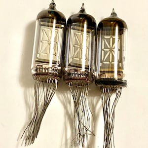 Może przedstawiać: Trzy zabytkowe lampy Nixie z szklanymi obudowami i metalowymi pinami. Każda lampa ma ciemną, zaokrągloną górę i wyświetla segmentowy wyświetlacz cyfrowy. Rurki są ułożone pionowo na białym tle, prezentując swój unikalny design.
