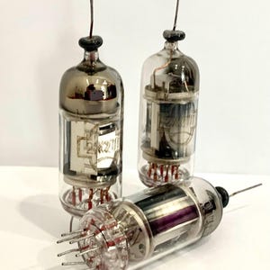 Op de afbeelding: Een verzameling vintage vacuümbuizen met heldere glazen lichamen, metalen toppen en meerdere pinnen. De buizen hebben interne componenten zichtbaar door het glas, waarbij sommige tekst weergeven. Eén buis ligt horizontaal, de andere staan rechtop.