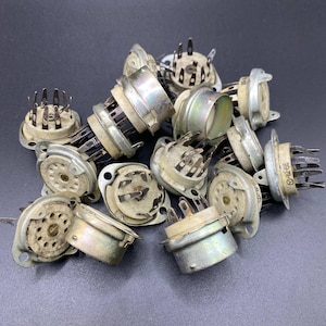 May include: A collection of vintage electronic vacuum tube sockets featuring circular off-white ceramic bases with metallic silver-toned cylindrical housings and multiple protruding metal contact pins arranged in a circular pattern for electrical connections.