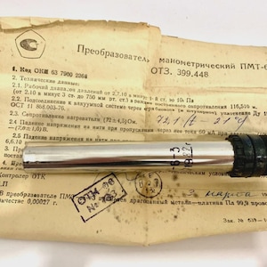 Pode incluir: Um transdutor de pressão vintage, modelo PMT-6-3, com corpo prateado e conector preto. O dispositivo repousa sobre um documento com especificações técnicas, selos e anotações manuscritas. O documento está amarelado.
