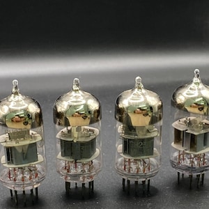 Pode incluir: Quatro tubos de vácuo de vidro transparente dispostos em fila, cada um com uma cúpula metálica superior, componentes internos de metal cinza e uma base circular com vários pinos metálicos finos para conexão elétrica e integração de circuitos.