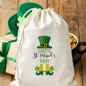 Puede incluir: Una bolsa blanca con cordón con un sombrero verde de duende y el texto "Happy St. Patrick's Day!". Un sombrero verde, una caja de regalo y una taza negra con monedas de oro están en el fondo.