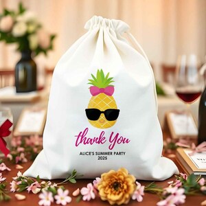 May include: A white drawstring bag featuring a cartoon pineapple wearing sunglasses and a pink bow. The text "thank You" is written in pink script, with "ALICE'S SUMMER PARTY 2025" below. The bag is on a table with flowers.