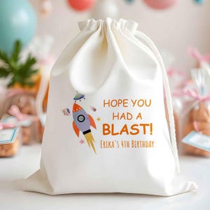 May include: Off-white drawstring bag with a colorful rocket ship graphic and the text "HOPE YOU HAD A BLAST!" and "ERIKA'S 4TH BIRTHDAY" in orange. The bag is likely for a birthday party.