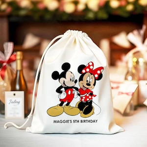 May include: A cream-coloured drawstring bag with a cartoon illustration of Mickey and Minnie Mouse. Mickey wears red shorts, and Minnie a red polka dot dress. The text "MAGGIE'S 5TH BIRTHDAY" is printed below the characters.