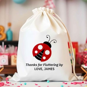 May include: A white drawstring bag featuring a red ladybug design with black accents. The bag has the text "Thanks for Fluttering by LOVE, JAMES" printed in black. The bag is made of a soft, opaque material.