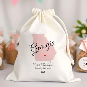 May include: A white drawstring bag featuring a pink outline of the state of Georgia with the word "Georgia" in elegant script. Below, it reads "Carter Randolph Family Reunion 2025". A small red heart is also present.