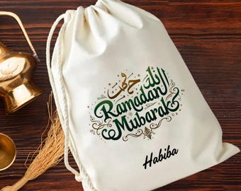 May include: A white drawstring bag with the words "Ramadan Mubarak" in green and gold script. The name "Habiba" is printed in black below the text. The bag is made of a soft fabric and has a white drawstring closure.