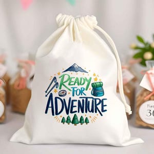 May include: A cream-coloured drawstring bag featuring the phrase "Ready for Adventure" in blue and green. The design includes a mountain, compass, backpack, and trees. The bag is made of a soft, opaque material.
