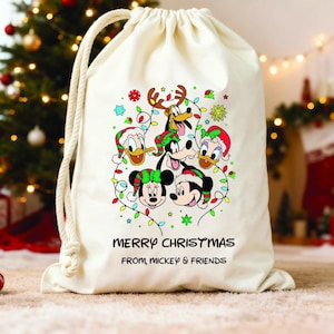 Könnte beinhalten: Ein cremefarbener Kordelzugbeutel mit einer farbenfrohen Grafik von Mickey Mouse, Minnie Mouse, Donald Duck und Goofy, die Weihnachtsmützen tragen und von Lichterketten umgeben sind. Der Text "MERRY CHRISTMAS FROM, MICKEY & FRIENDS" ist darunter gedruckt.