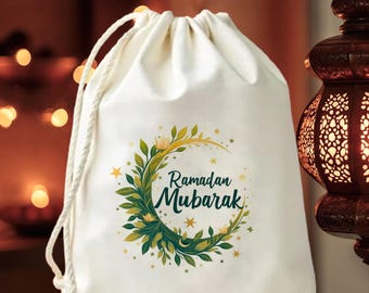 May include: A white drawstring bag with the words "Ramadan Mubarak" in green and black script, surrounded by a crescent moon and floral design. The name "Khalid" is printed below. The bag has a white rope drawstring.