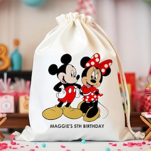 May include: A white drawstring bag with Mickey and Minnie Mouse illustrations. Mickey wears red shorts, and Minnie a red polka dot dress. The text "MAGGIE'S 5TH BIRTHDAY" is printed below the characters.