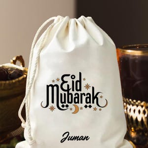May include: A cream-coloured drawstring bag featuring the words "Eid Mubarak" in black and gold script, decorated with stars and a crescent moon. The name "Juman" is printed below.