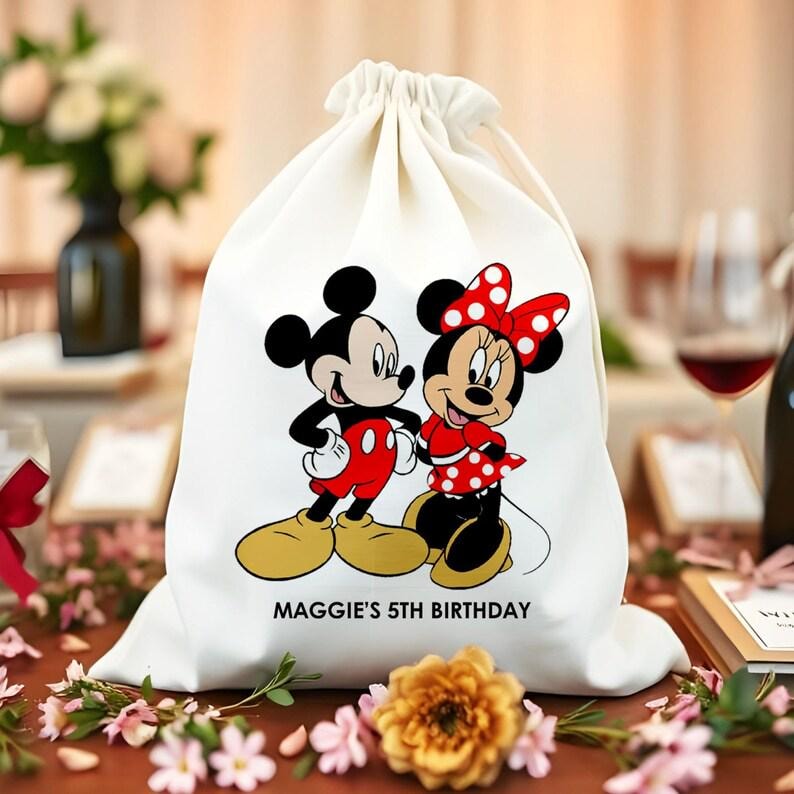May include: A white drawstring bag with Mickey and Minnie Mouse printed on it. Mickey wears red shorts and Minnie a red polka dot dress. The text "MAGGIE'S 5TH BIRTHDAY" is below the characters. The bag is on a brown surface.