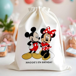 May include: A cream-coloured drawstring bag with a cartoon illustration of Mickey and Minnie Mouse. Minnie wears a red dress with white polka dots and a matching bow. The text "MAGGIE'S 5TH BIRTHDAY" is printed below the characters.