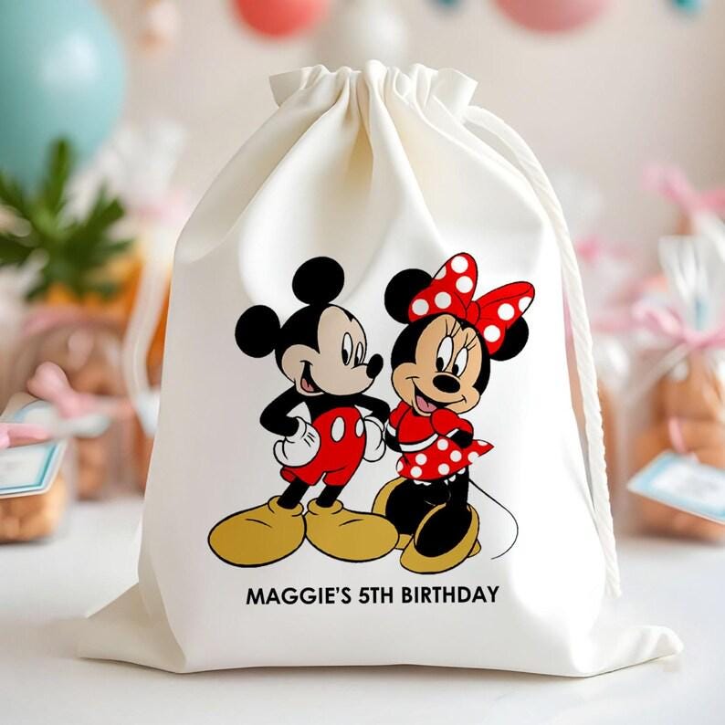 May include: A cream-coloured drawstring bag with a cartoon illustration of Mickey and Minnie Mouse. Minnie wears a red dress with white polka dots and a matching bow. The text "MAGGIE'S 5TH BIRTHDAY" is printed below the characters.