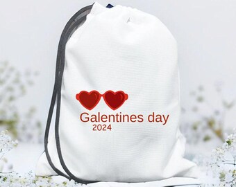 Może przedstawiać: Biała torba ze sznurkiem z czerwonymi okularami przeciwsłonecznymi w kształcie serca i napisem "Galentines day 2024". Torba ma ciemnoszary sznurek. Idealna do przenoszenia prezentów lub niezbędnych rzeczy.
