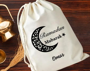 May include: A white drawstring bag with a black crescent moon design and the words "Ramadan Mubarak" and the name "Omar". The bag is made of a soft fabric and has a drawstring closure.