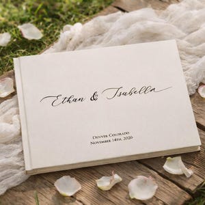 以下が含まれることがあります： 「Ethan & Isabella」の名前がエレガントな筆記体で書かれたクリーム色のウェディングアルバム。アルバムは、花びらが散らばり、柔らかい白い布が敷かれた木製の表面に置かれています。「Denver Colorado November 14th, 2026」の文字も表示されています。