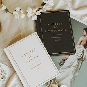 Op de afbeelding: Een bovenaanzicht met huwelijksthema-items. Twee boeken met gouden letters lezen "A Letter to My Husband" en "A Letter to My Wife". Een tijdschrift met een bruid in een trouwjurk is zichtbaar. Een gouden kandelaar en bloemenkrans maken de scène compleet.