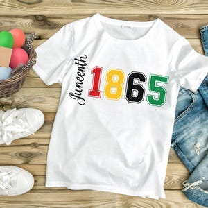 Pode incluir: Camiseta branca com a palavra "Juneteenth" em escrita preta e os números "1865" em vermelho, amarelo, preto e verde. A camiseta está em uma superfície de madeira, com tênis brancos e uma cesta de ovos coloridos.