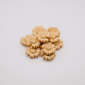 May include: A cluster of light tan, flower-shaped cookies. Each cookie has a detailed floral design with a central disc and radiating petals. The cookies are arranged on a white background.