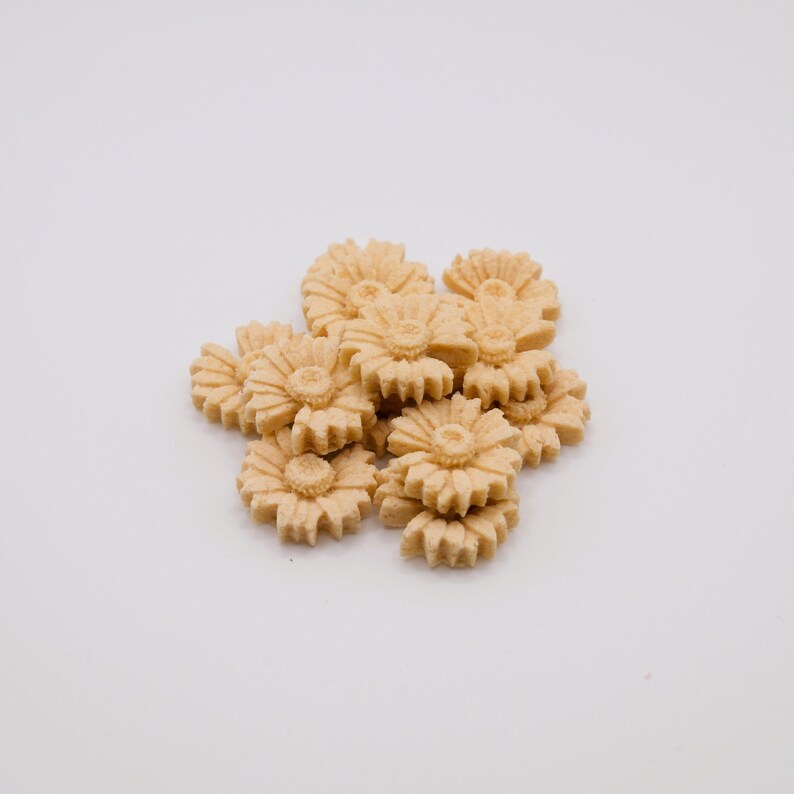 May include: A cluster of light tan, flower-shaped cookies. Each cookie has a detailed floral design with a central disc and radiating petals. The cookies are arranged on a white background.