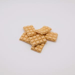 May include: A pile of small, square, light brown waffles with a grid pattern. The waffles are stacked on a white background, and the image is well-lit, highlighting the texture and color of the baked goods.