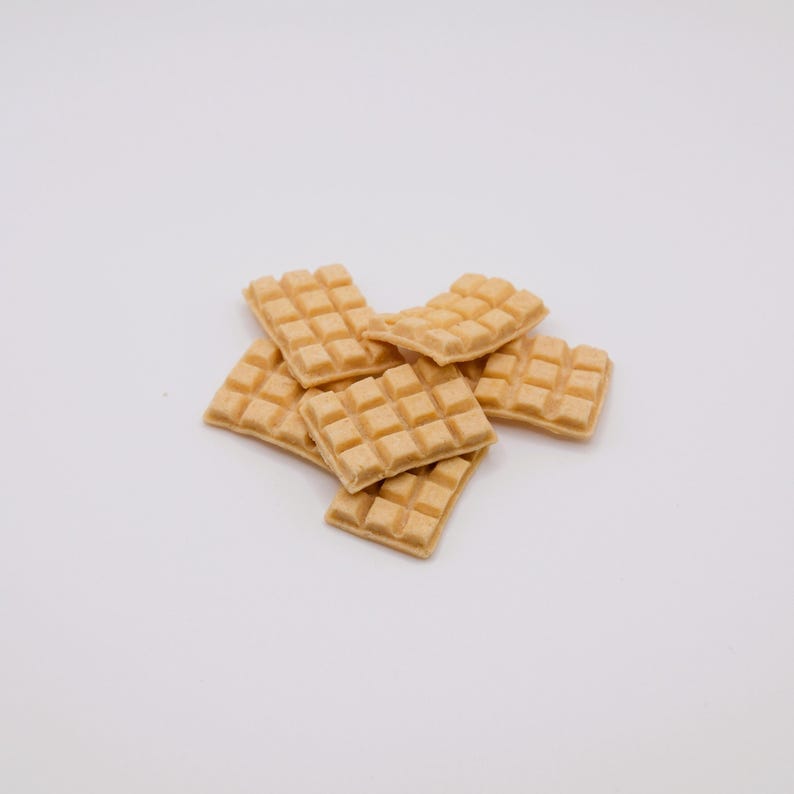 May include: A pile of small, square, light brown waffles with a grid pattern. The waffles are stacked on a white background, and the image is well-lit, highlighting the texture and color of the baked goods.