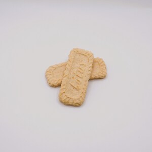 May include: Two rectangular, light brown cookies with a textured border and a central design. One cookie is positioned atop another, with the top cookie displaying a visible pattern. The cookies are set against a plain white background.