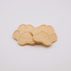 May include: Two light brown, paw-shaped cookies, each with five circular indentations representing the pads and toes. The cookies are set against a plain white background, suggesting a pet-themed treat or craft item.