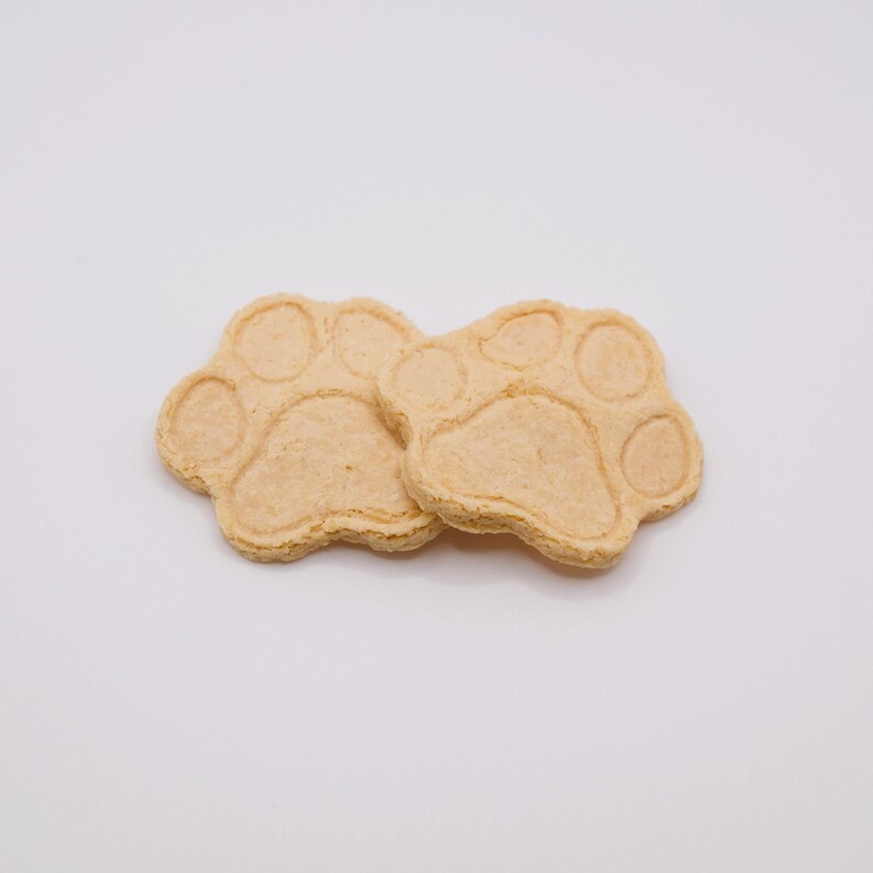 May include: Two light brown, paw-shaped cookies, each with five circular indentations representing the pads and toes. The cookies are set against a plain white background, suggesting a pet-themed treat or craft item.