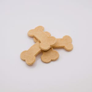 May include: A pile of tan, bone-shaped dog treats on a white background. The treats have a slightly textured surface and are arranged in a casual, overlapping manner. The treats are a light brown color.
